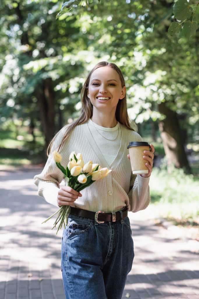 cheerful woman in glasses and jeans walking with tulips and paper cup in park - Fotoğraf, Görsel