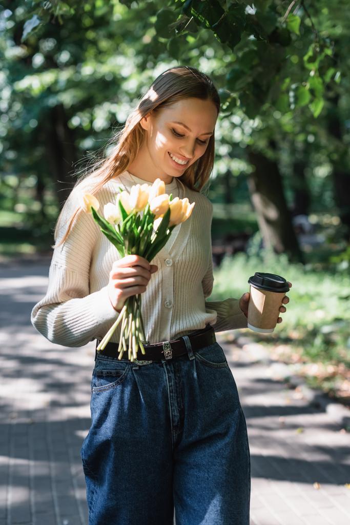 happy woman in blue denim jeans walking with bouquet of tulips and paper cup in green park - Fotoğraf, Görsel