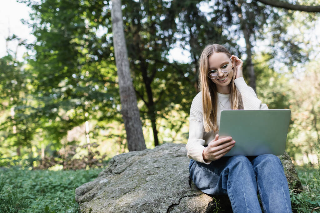 low angle view of happy woman in glasses adjusting hair and looking at laptop in park - Foto, immagini