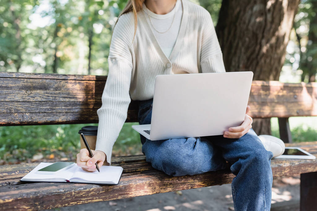 vista cortada da mulher usando laptop enquanto escreve em notebook perto de gadgets no banco - Foto, Imagem