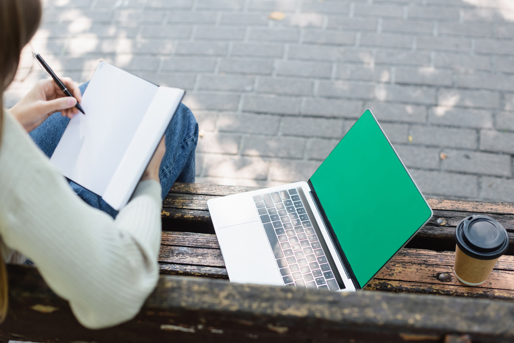 Вид сверху женщины, пишущей в тетрадке рядом с ноутбуком с зеленым экраном и бумажной чашкой  - Фото, изображение
