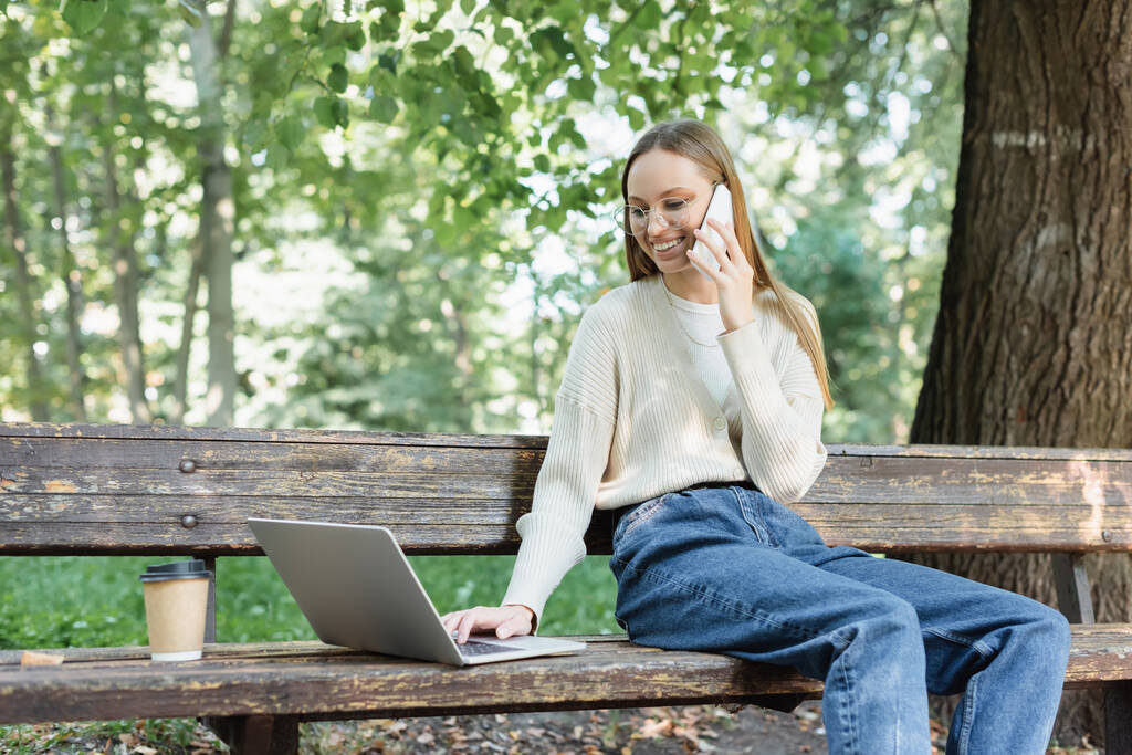 cheerful woman in glasses talking on smartphone while using laptop on bench - 写真・画像