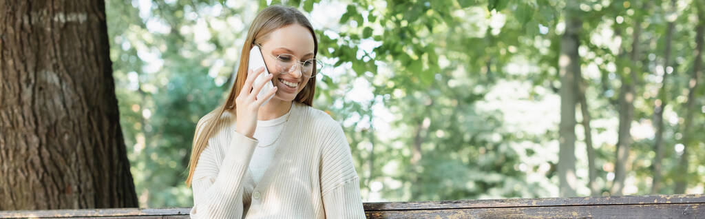 веселая женщина в очках разговаривает по смартфону в зеленом парке, баннер - Фото, изображение
