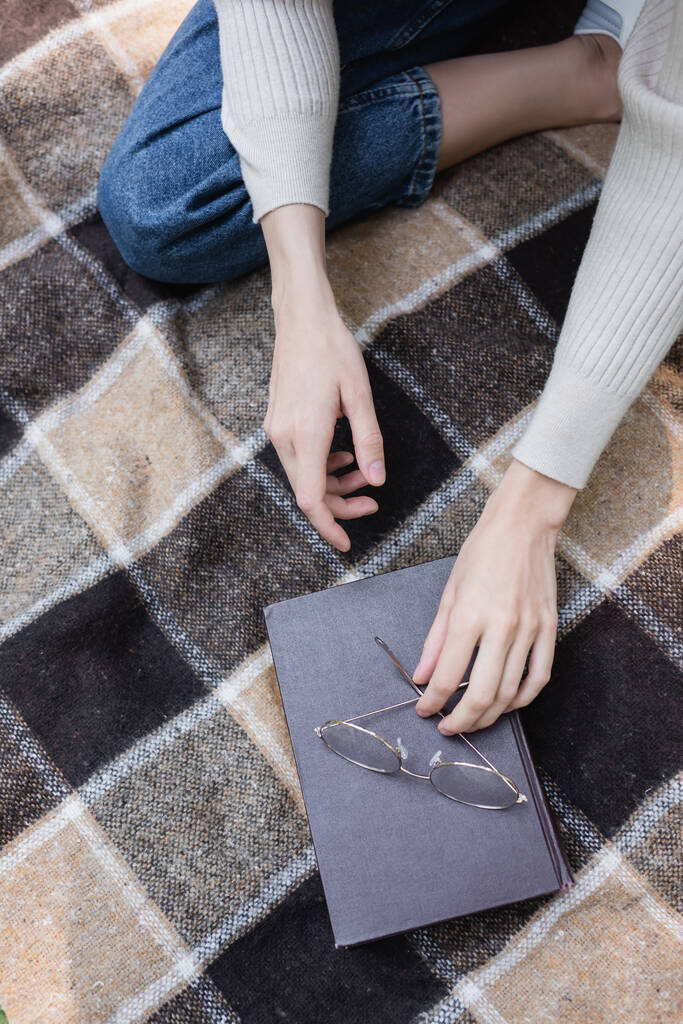 top view of woman reaching glasses on book while sitting on checkered blanket  - Photo, Image