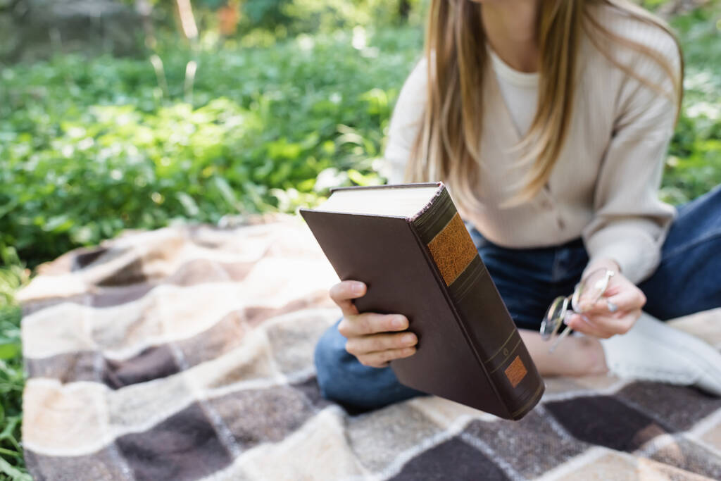 cropped view of woman holding book and glasses while sitting on checkered blanket  - Photo, image