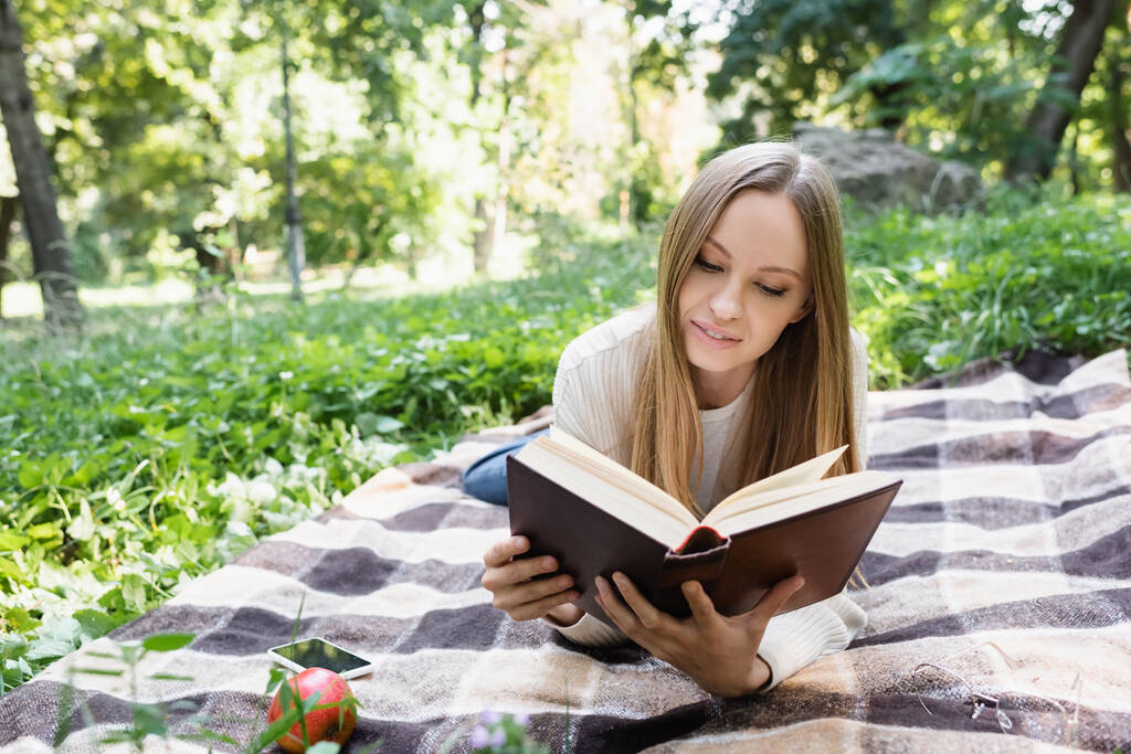 mulher lendo livro enquanto deitado perto de smartphone e maçã no cobertor  - Foto, Imagem
