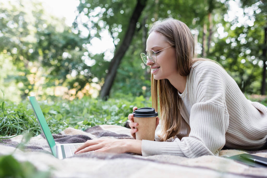 happy freelancer in glasses holding paper cup while lying on blanket and using laptop in park  - Фото, изображение