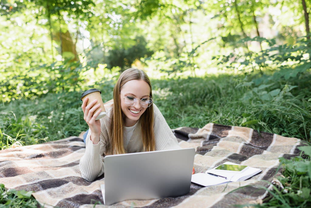 usmívající se žena v brýlích ležící na přikrývce, držící papírový šálek a používající notebook v parku  - Fotografie, Obrázek