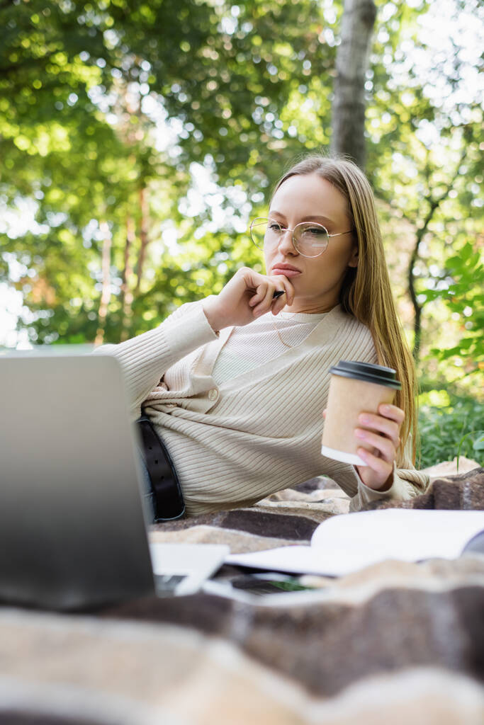 σκεπτική γυναίκα σε γυαλιά κρατώντας χάρτινο κύπελλο και κοιτάζοντας φορητό υπολογιστή σε κουβέρτα στο πάρκο - Φωτογραφία, εικόνα