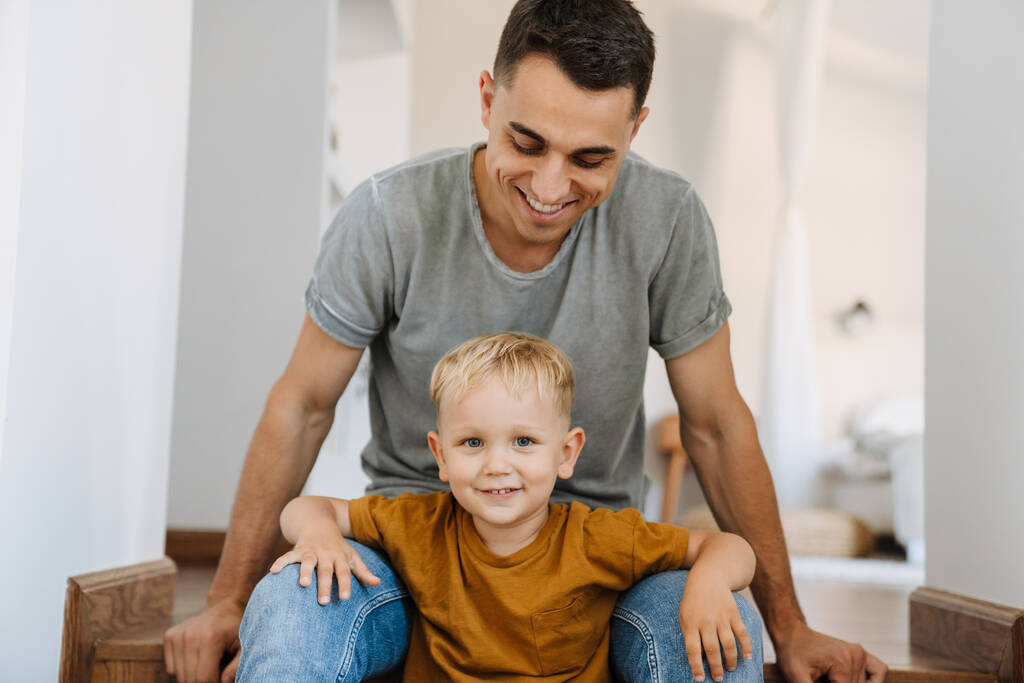 Mladý bílý otec se usmívá, zatímco sedí se svým synem doma na schodech - Fotografie, Obrázek