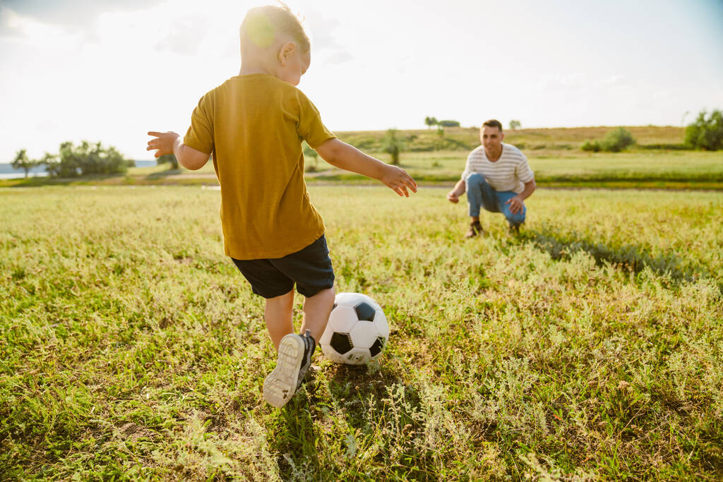 Ο νεαρός λευκός πατέρας και ο μικρός γιος του παίζουν ποδόσφαιρο έξω. - Φωτογραφία, εικόνα