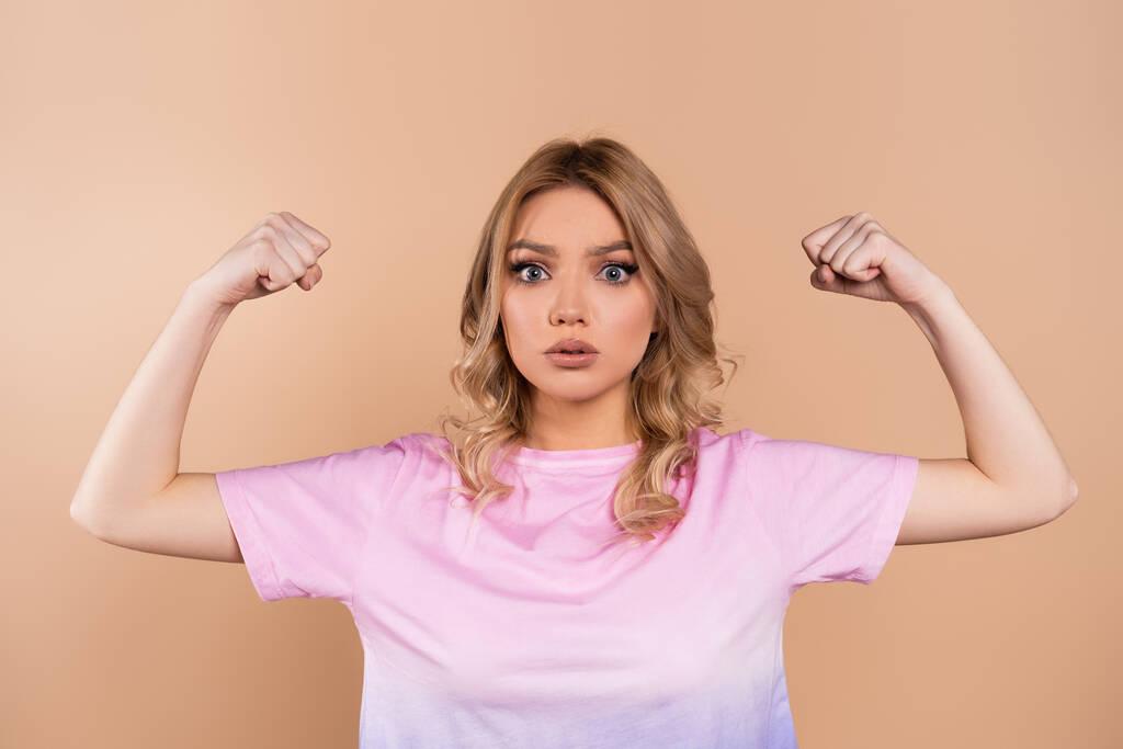 jonge vrouw tonen spieren en kijken naar camera geïsoleerd op beige - Foto, afbeelding