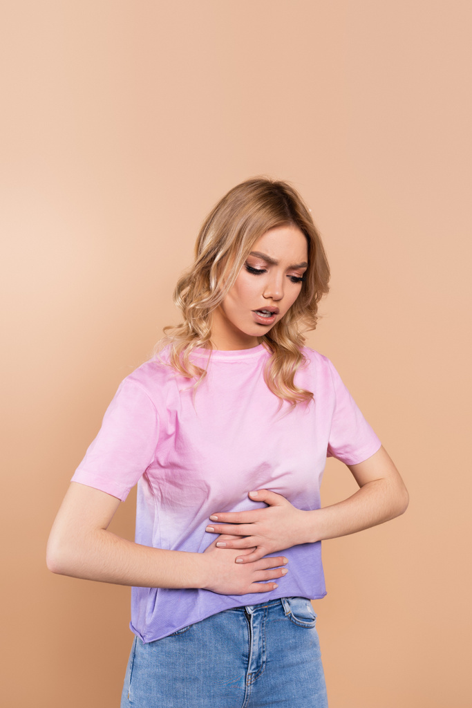 displeased woman touching belly while suffering from abdominal pain isolated on beige - Фото, изображение