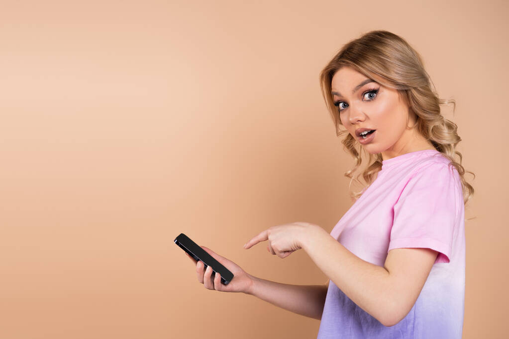 amazed woman pointing at mobile phone and looking at camera isolated on beige - Foto, imagen