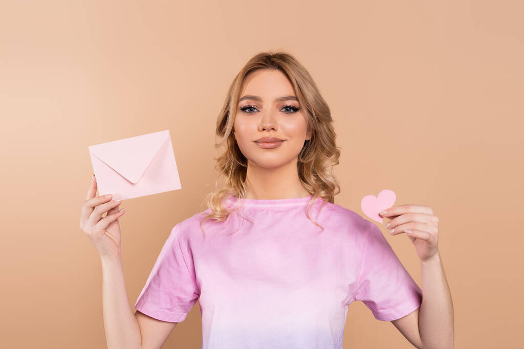 happy woman with wavy hair holding envelope and paper heart isolated on beige - Foto, afbeelding