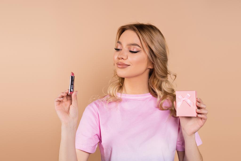 pleased woman holding lipstick and small gift box isolated on beige - Fotografie, Obrázek