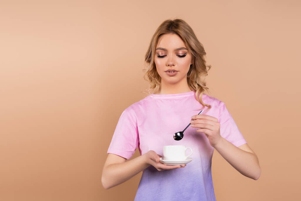 Dalgalı saçlı, elinde kahve fincanı ve bej rengi çay kaşığı tutan genç bir kadın. - Fotoğraf, Görsel