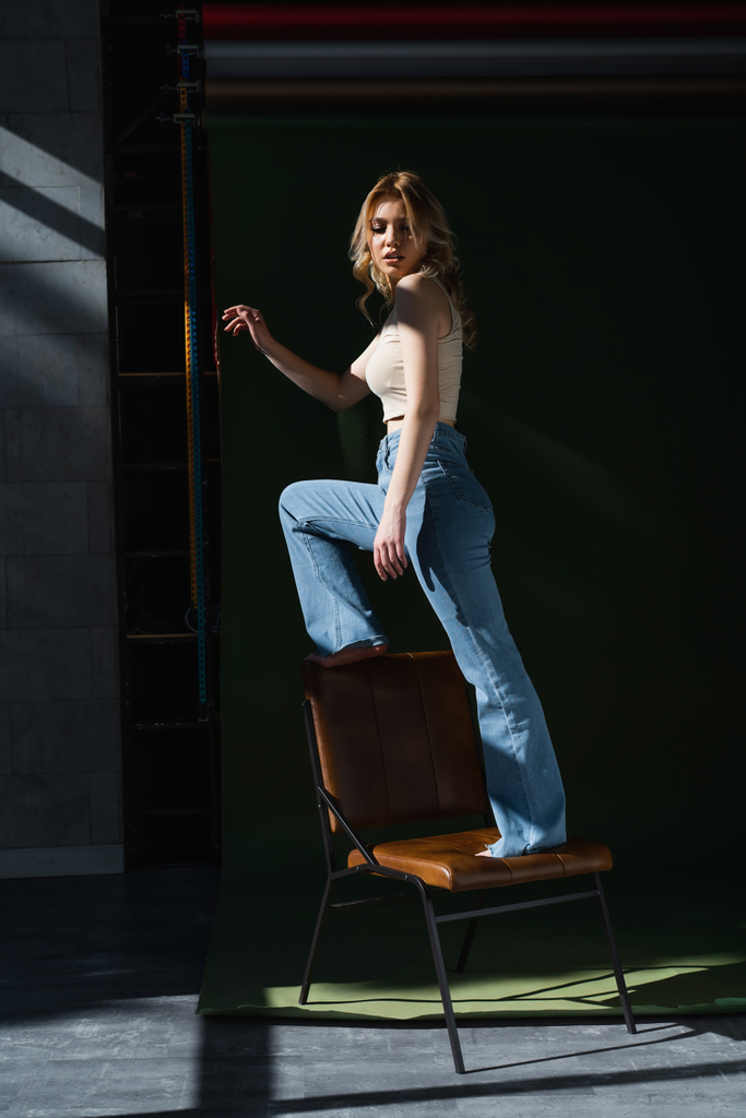full length view of slim woman in jeans standing on chair on dark background - Photo, Image