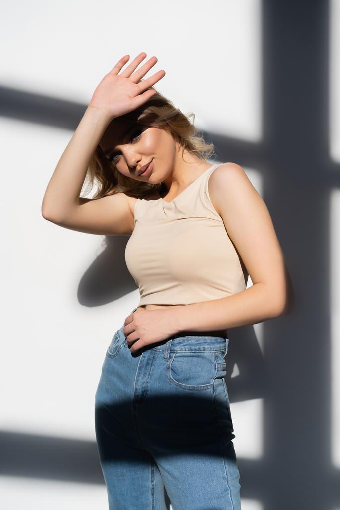 young woman in jeans looking at camera from under her arm on white background with shadows - Photo, Image