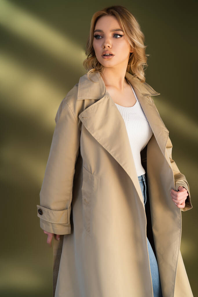 woman with wavy hair looking away while posing in trench coat on beige background - Photo, image