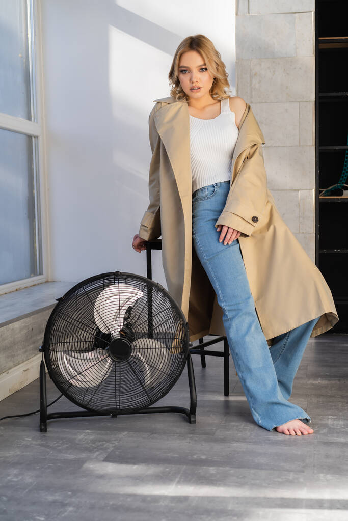 full length view of sensual barefoot woman in jeans and trench coat posing near huge electric fan - 写真・画像