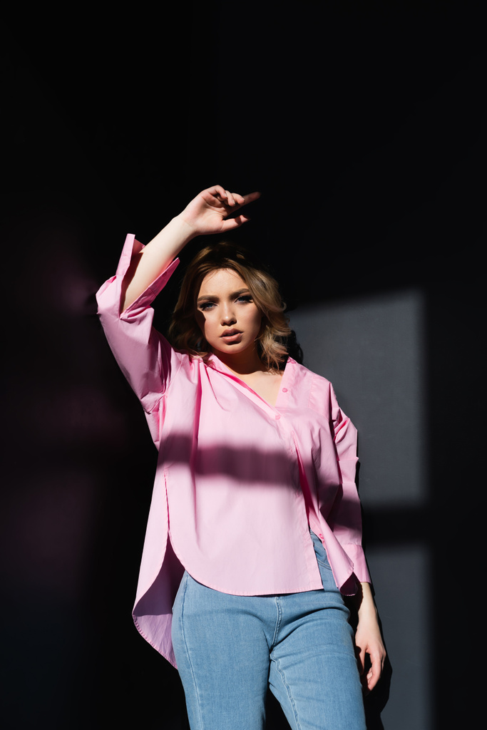 young woman in pink shirt standing in lighting with raised hand on dark background - Foto, afbeelding