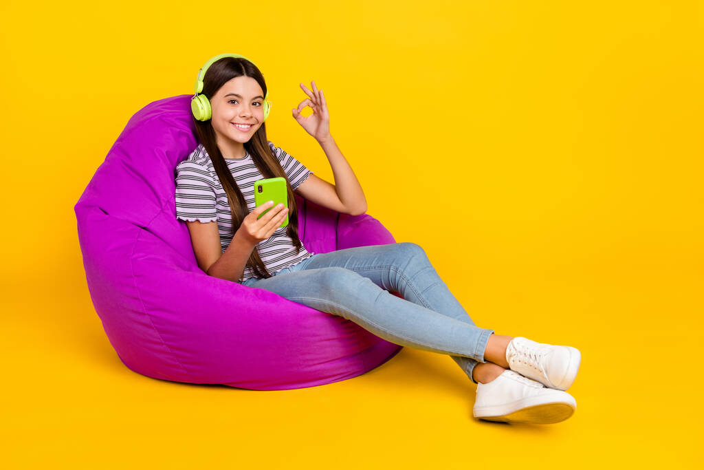 Full size profile side photo of young girl sit beanbag show fingers okey symbol use mobile isolated over yellow color background - Foto, Imagem