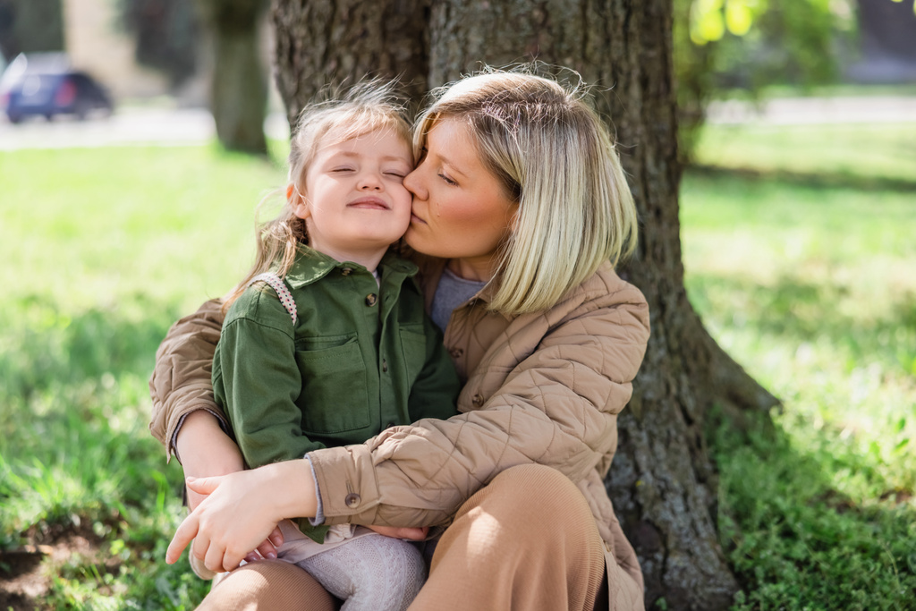 woman kissing happy daughter while sitting under trees in park - Photo, image