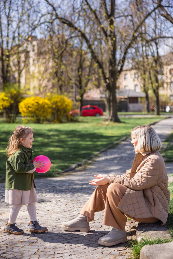 oldal kilátás vidám lány holding ball játék közben anya a parkban - Fotó, kép