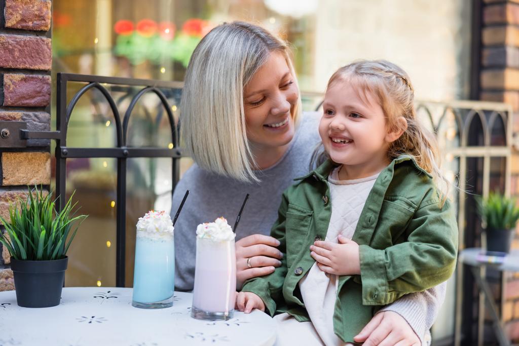 blij meisje aanraken buik in de buurt bril met heerlijke milkshake en mam - Foto, afbeelding