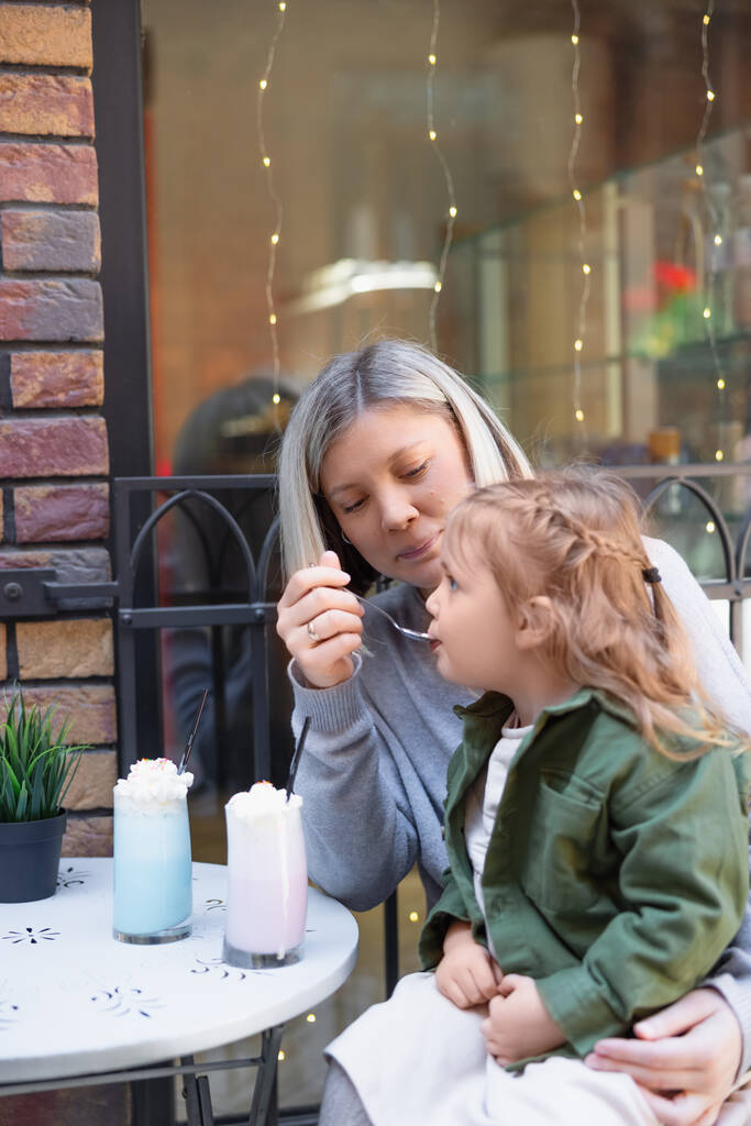 happy woman feeding daughter with milk dessert in cafe on urban street - Zdjęcie, obraz