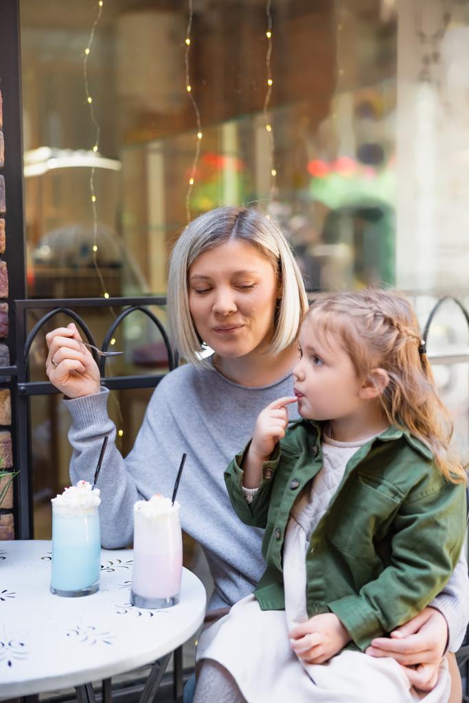 smiling woman holding spoon near daughter and tasty milkshake in street cafe - Photo, Image
