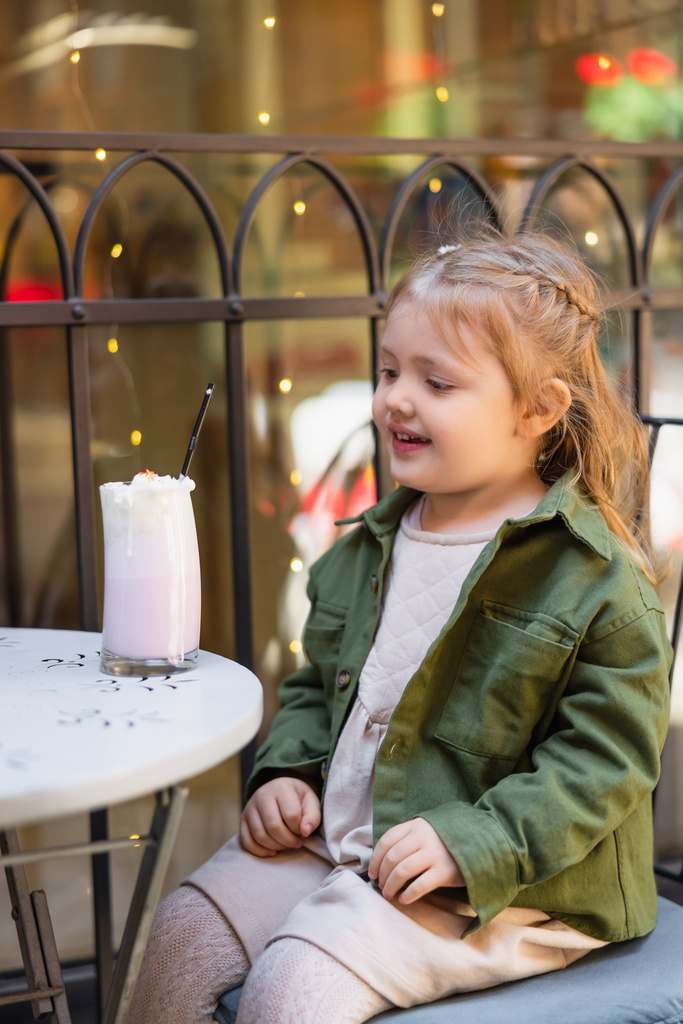 smiling child looking at glass with milkshake while sitting in street cafe - Photo, image