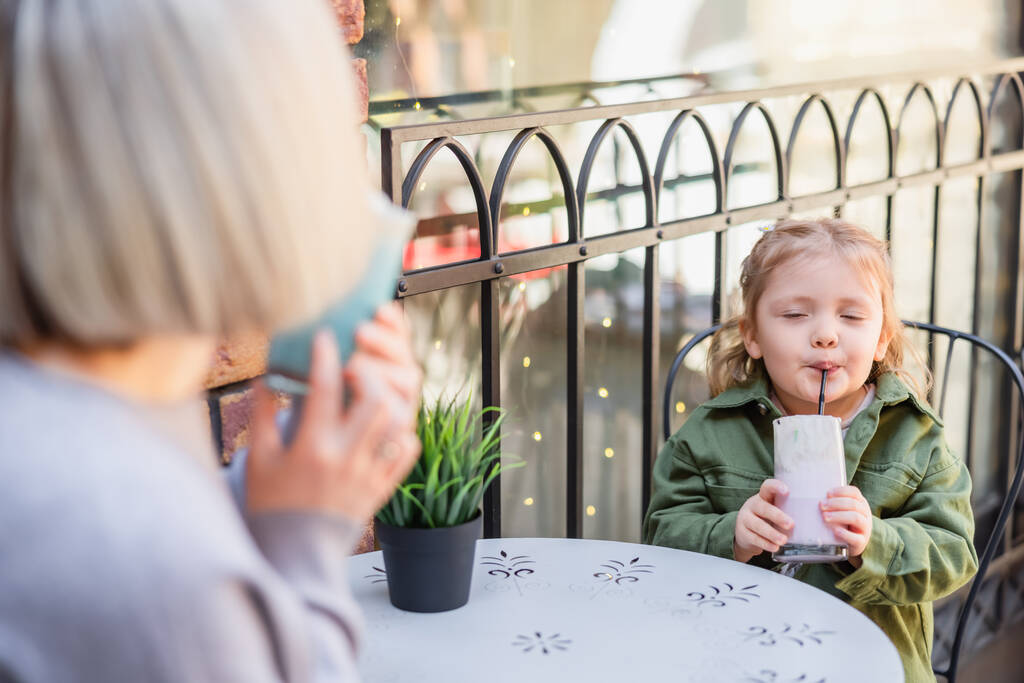 chica con los ojos cerrados disfrutando de delicioso postre cerca borrosa mamá en la cafetería en la calle - Foto, imagen