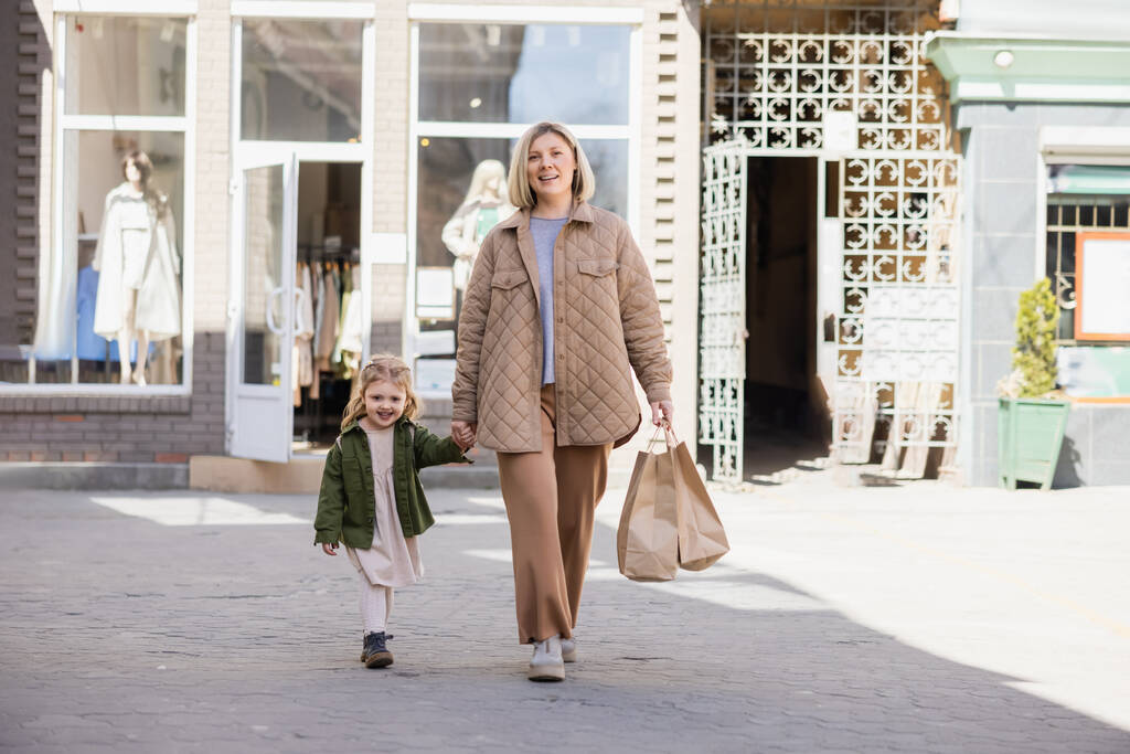 mulher sorrindo com sacos de compras olhando para a câmera enquanto caminhava com a criança na rua - Foto, Imagem