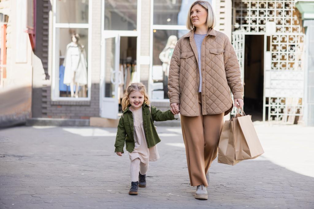 trendige Frau mit Einkaufstaschen und fröhlichem Mädchen, das Händchen hält, während es auf der städtischen Straße läuft - Foto, Bild
