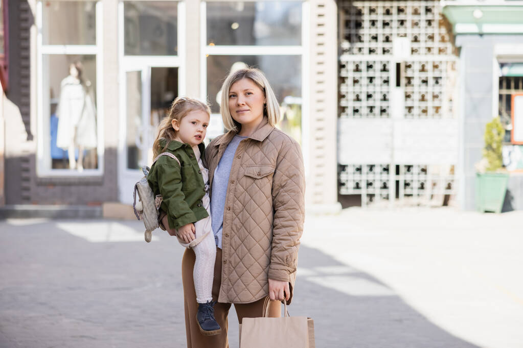 positive and trendy woman with shopping bags looking away while holding daughter outdoors - Foto, Imagen