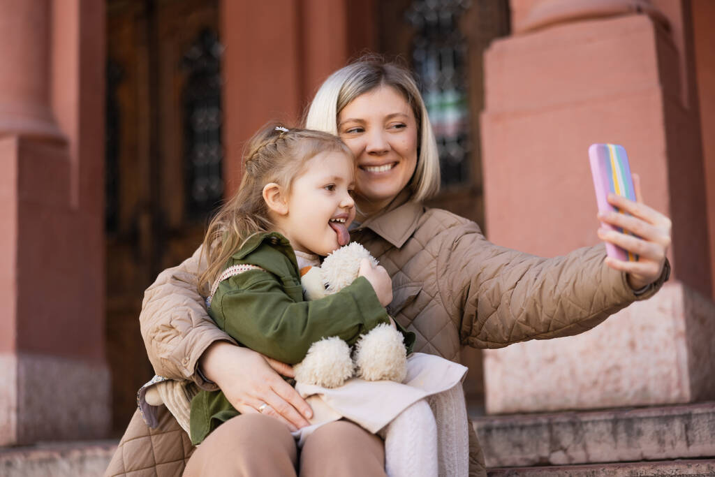 little girl with soft toy sticking out tongue near smiling mother taking selfie on mobile phone - Фото, изображение