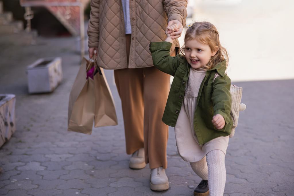 excited child holding hands with mom walking with shopping bags outdoors - Zdjęcie, obraz