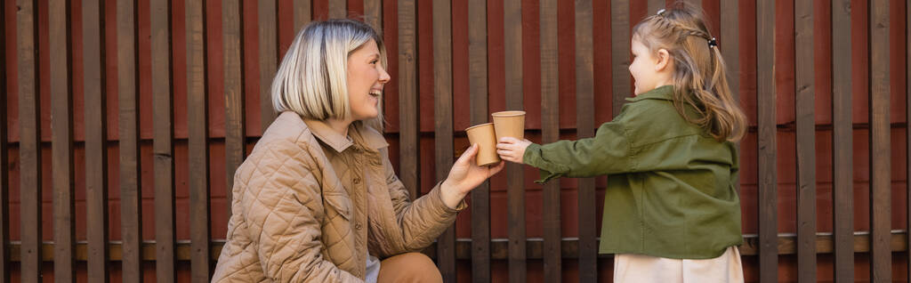 side view of cheerful woman and girl toasting with paper cups near wooden fence outdoors, banner - Foto, Imagem