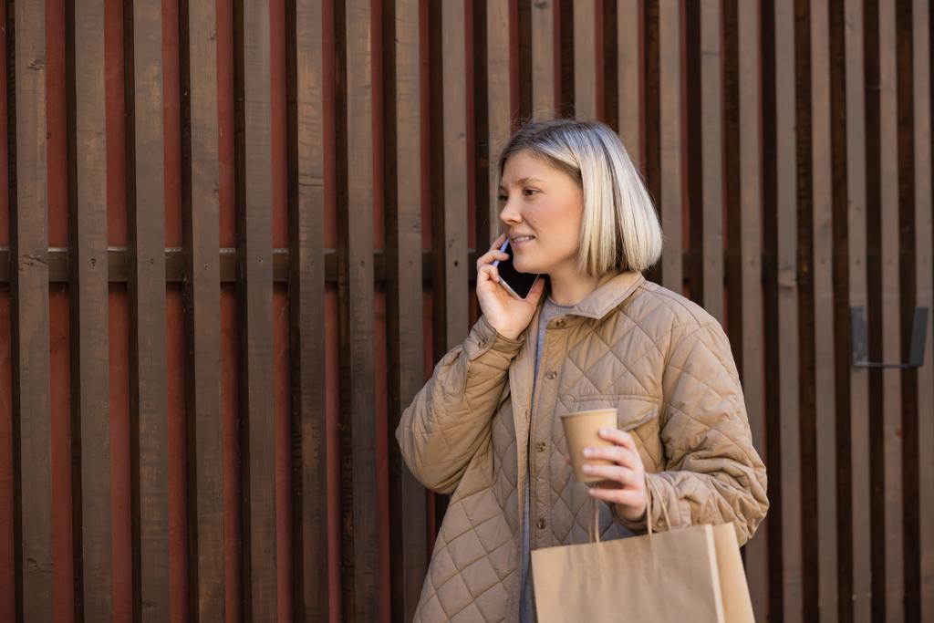mujer rubia sonriente con café para ir y bolsa de compras hablando en el teléfono inteligente cerca de valla - Foto, Imagen