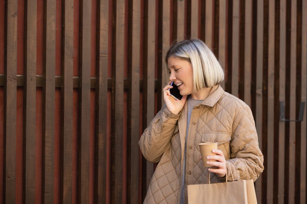 ευτυχισμένη γυναίκα σε μπεζ σακάκι μιλάμε στο κινητό τηλέφωνο, ενώ κρατώντας χάρτινο κύπελλο και τσάντα ψώνια - Φωτογραφία, εικόνα