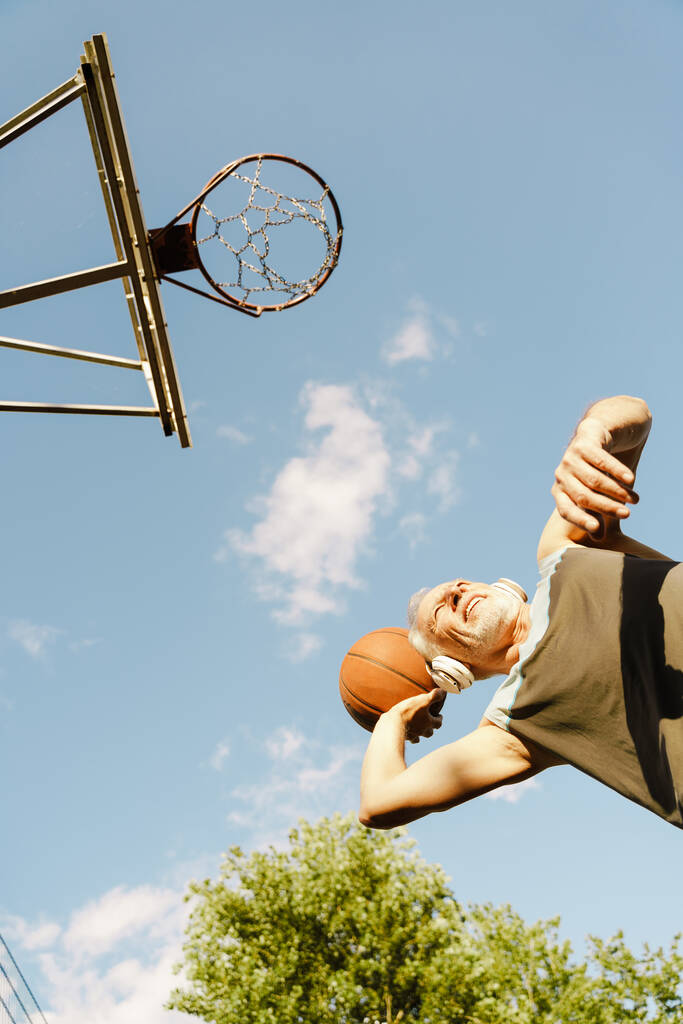 Harmaa vanhempi mies kuuntelee musiikkia pelatessaan koripalloa urheilukentällä - Valokuva, kuva