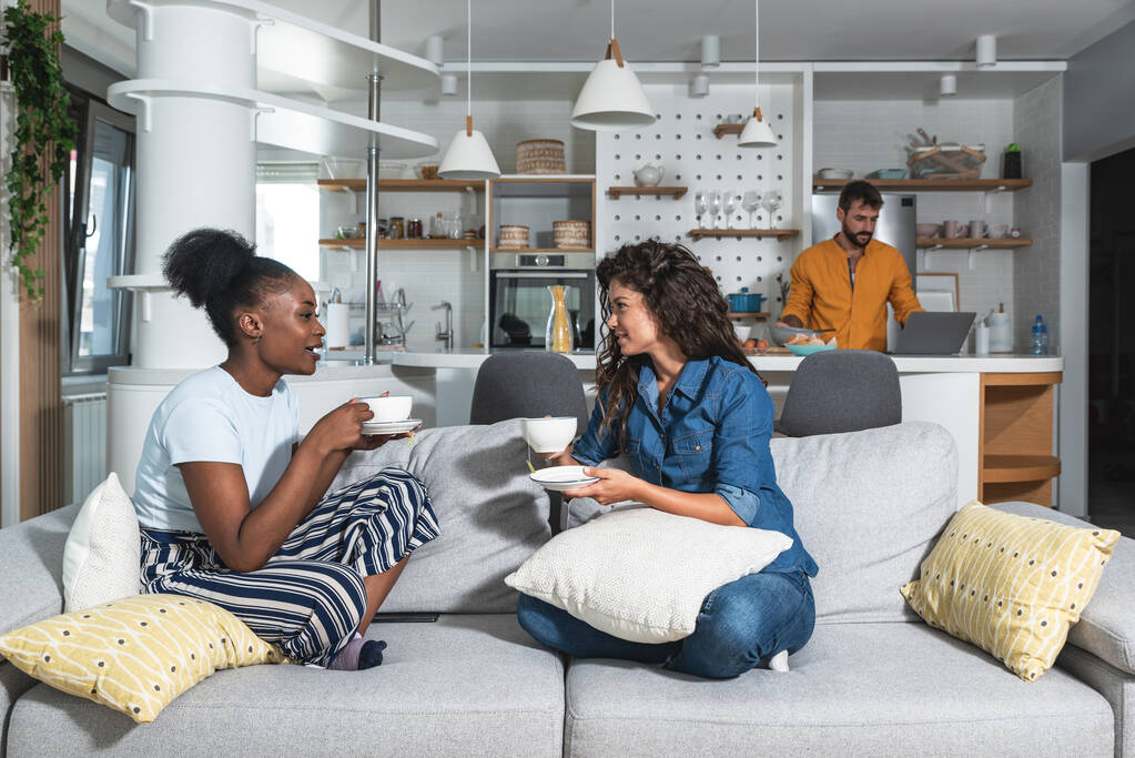 2人の若い女性はソファに座ってお茶やコーヒーを飲みながら女性の問題について話している間、男は台所に立って、彼らのために昼食を調理する彼らのためにラップトップ上のインターネットからレシピを読む - 写真・画像