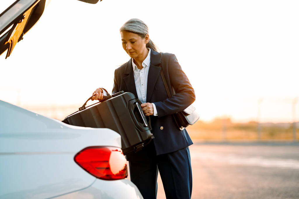 Ώριμη Ασιάτισσα επιχειρηματίας που φοράει κοστούμι και βάζει τη βαλίτσα της στο πορτ-μπαγκάζ - Φωτογραφία, εικόνα