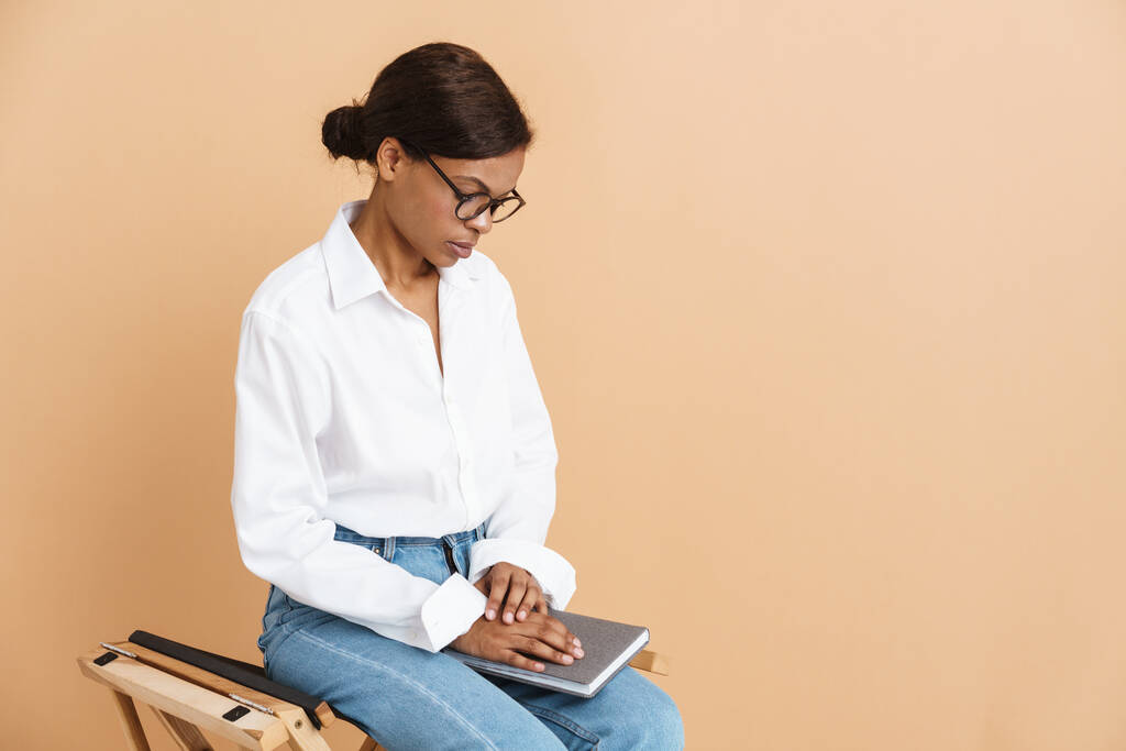 Νεαρή μαύρη γυναίκα που κάθεται με σχεδιαστή σε καρέκλα απομονωμένη πάνω από μπεζ φόντο - Φωτογραφία, εικόνα