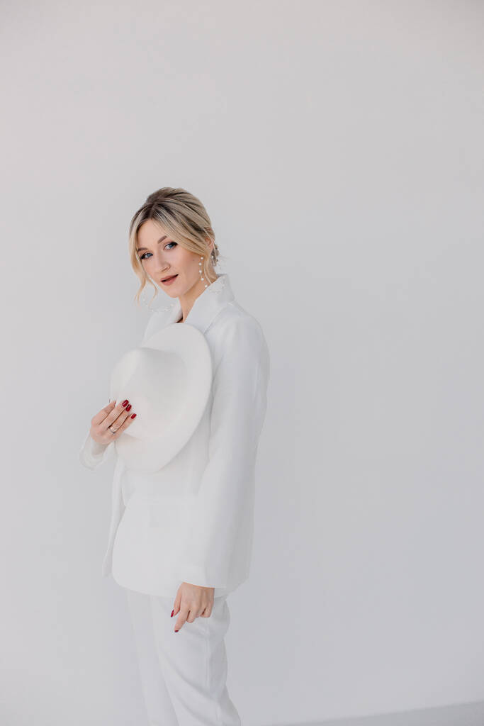 Confident young blond woman smiling, looking at camera isolated on white background. Studio portrait of successful friendly female in white suit and hat, posing over white wall. - Photo, Image