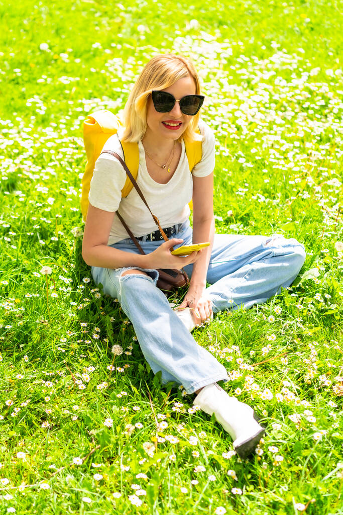 Portrait of a blonde tourist woman with a hat and sunglasses sitting on the grass in spring next to daisies, looking at what to visit or maps in the city on the mobile - Фото, изображение