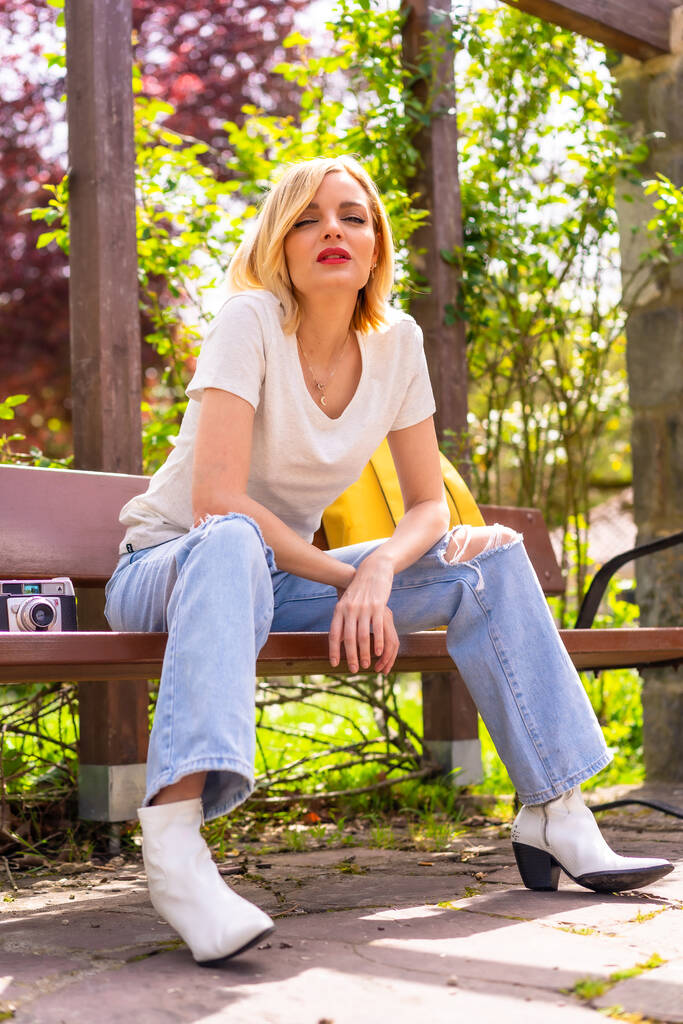 Portrait of a blonde tourist girl on summer vacation sitting on a bench in a park - 写真・画像