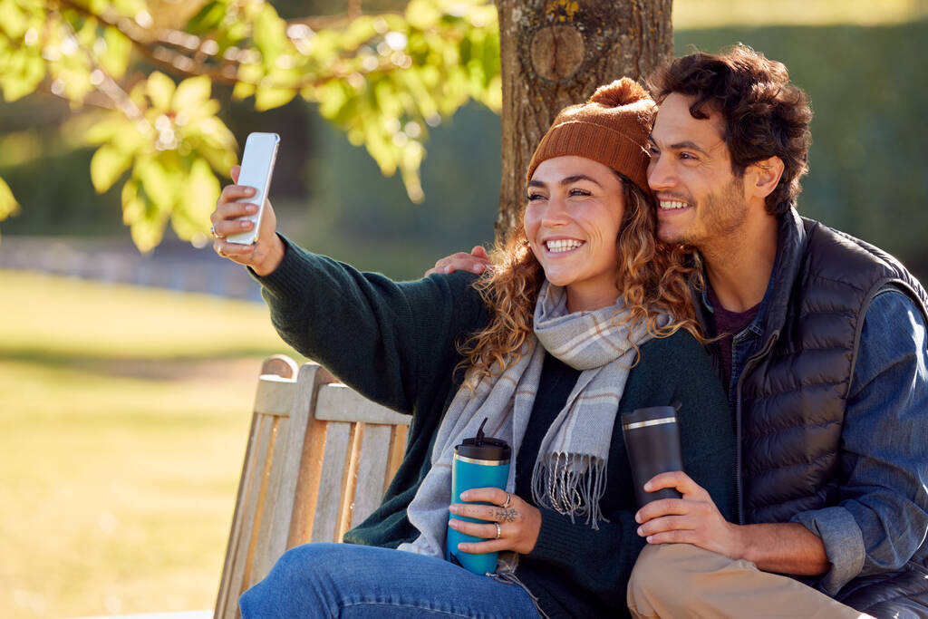 glimlachend paar nemen selfie met mobiele telefoon zitten op de bank in de herfst Park - Foto, afbeelding